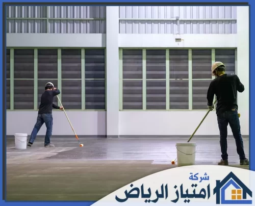 ايبوكسي رخام بالرياض 0506986916 شركة امتياز الرياض عاملان يرتديان خوذات أمان يضعان طبقة من الإيبوكسي العاكسة للضوء على أرضية جراج تجاري.